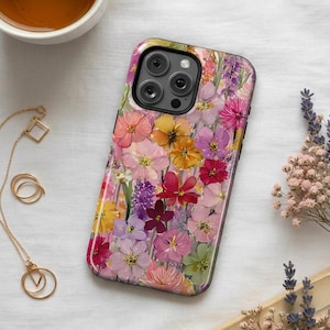 May include: A phone case adorned with a colourful floral pattern, showcasing a variety of flowers in pinks, yellows, purples, and reds. The case has a glossy finish and a black camera lens aperture. The design presents a botanical theme with diverse flower types.