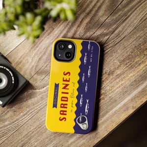 Phone case designed as a vintage sardine tin can with silver fish illustration and pull-tab detail on a bold yellow and navy background – retro foodie aesthetic.