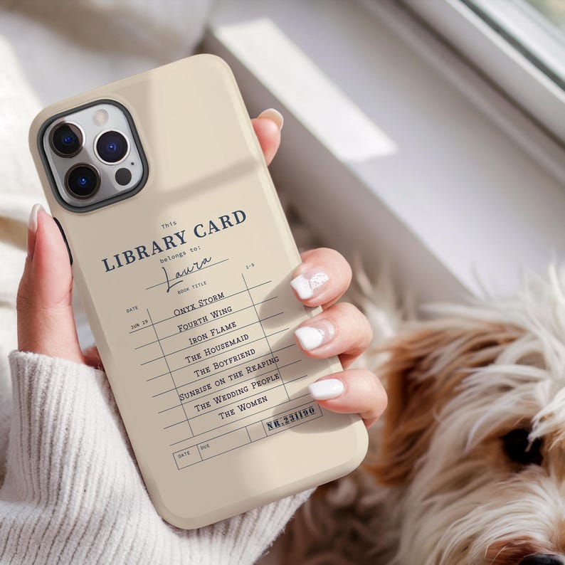 May include: A beige phone case featuring a vintage library card design. The text includes "LIBRARY CARD" and a list of book titles. The case is held in a hand, with a dog in the background.