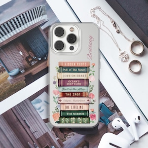 K&ouml;nnte beinhalten: Transparente Handyh&uuml;lle mit einem Design aus gestapelten B&uuml;chern, darunter Titel wie "Hidden Roots" und "Love on Heart". Der Name "Brittany" ist ebenfalls zu sehen. Eine silberne Halskette und Ringe liegen daneben.