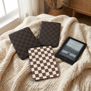 Puede incluir: Cuatro fundas para lectores de libros electrónicos Kindle. Tres tienen un patrón de cuadros marrones y negros, y una tiene un patrón de cuadros marrones y blancos. Un Kindle muestra el libro "The Beginning Life" de Brian Holer.