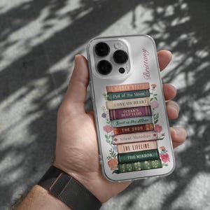 K&ouml;nnte beinhalten: Transparente Handyh&uuml;lle mit einem Stapel von B&uuml;chern, darunter Titel wie "Hidden Roots" und "Love on Heart." Die H&uuml;lle hat ein Blumenmuster und den Namen "Brittany." Das Handy wird in einer Hand gehalten.