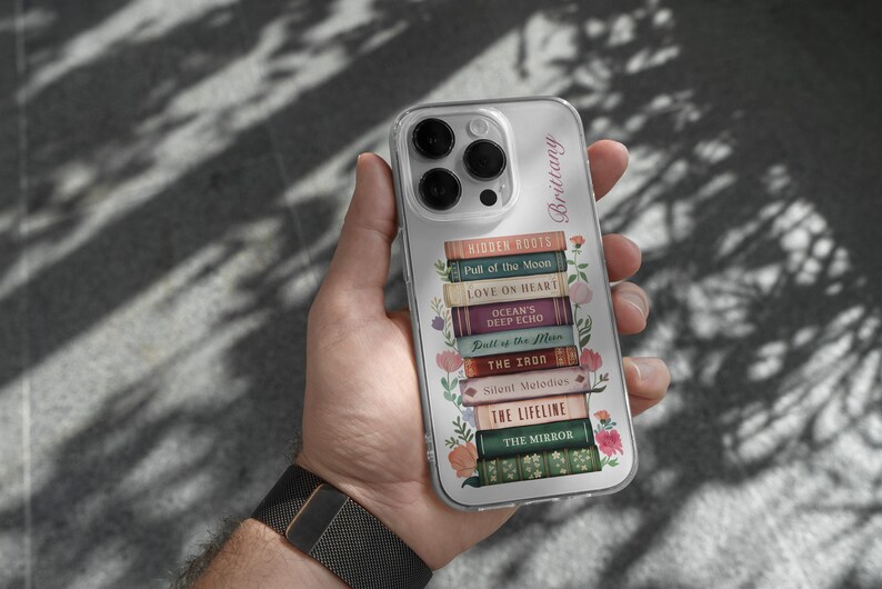K&ouml;nnte beinhalten: Transparente Handyh&uuml;lle mit einem Stapel von B&uuml;chern, darunter Titel wie "Hidden Roots" und "Love on Heart." Die H&uuml;lle hat ein Blumenmuster und den Namen "Brittany." Das Handy wird in einer Hand gehalten.