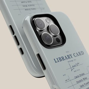 May include: A light blue phone case with a black camera ring and the text "This LIBRARY CARD belongs to: Laura". The case features a library card design with book titles and a date.