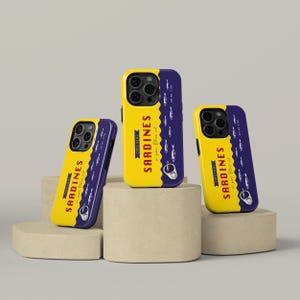 May include: Yellow and navy blue phone cases with a sardine can design. The cases feature the text "ORIGINAL SARDINES in pure olive oil" and a graphic of sardines. The cases are displayed on beige pedestals.