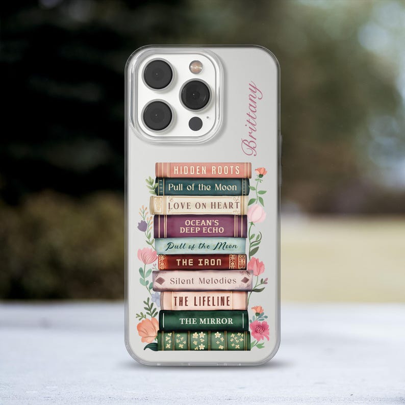 K&ouml;nnte beinhalten: Transparente Handyh&uuml;lle mit einem Stapel B&uuml;cher, darunter Titel wie "Hidden Roots" und "The Mirror." Blumendesigns und der Name "Brittany" sind aufgedruckt. F&uuml;r ein Smartphone.