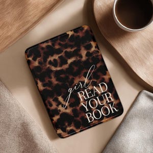 Op de afbeelding: Een rechthoekige e-reader hoes met een luipaardprint en de witte tekst "girl READ YOUR BOOK". De hoes is bruin en zwart. Een kopje koffie en een houten dienblad staan op de achtergrond.