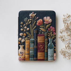 Puede incluir: Una funda rectangular para lector electrónico con cubierta de tela azul oscuro que presenta un bordado detallado de varios libros y jarrones llenos de flores en tonos rosa, amarillo y verde, creando una escena botánica texturizada.