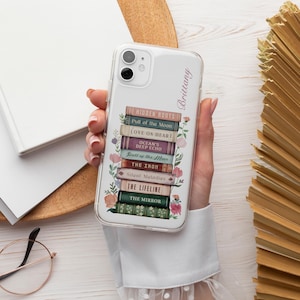 K&ouml;nnte beinhalten: Transparente Handyh&uuml;lle mit einem Design aus gestapelten B&uuml;chern. Die Titel lauten "Hidden Roots", "Pull of the Moon" und "Love on Heart". Blumendetails und der Name "Brittany" sind ebenfalls vorhanden. Die H&uuml;lle wird in einer Hand gehalten.