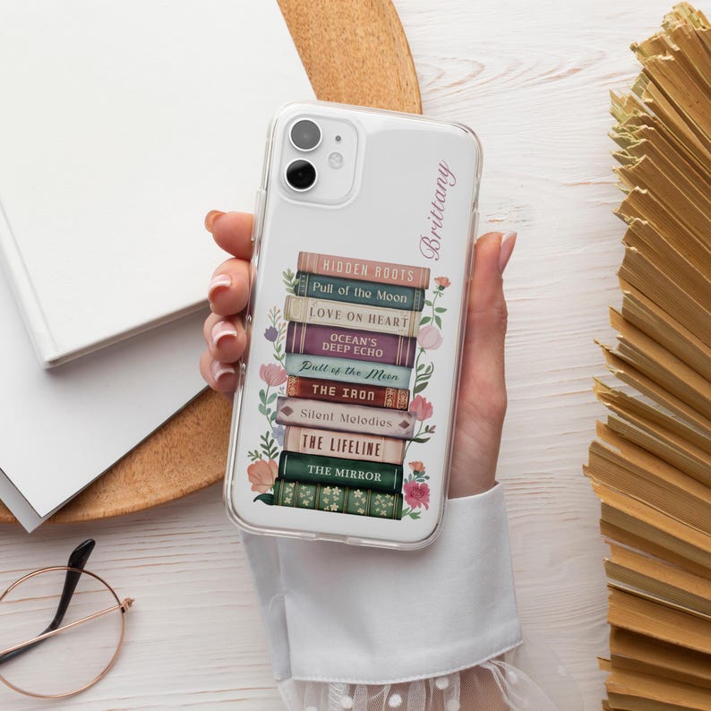 K&ouml;nnte beinhalten: Transparente Handyh&uuml;lle mit einem Design aus gestapelten B&uuml;chern. Die Titel lauten "Hidden Roots", "Pull of the Moon" und "Love on Heart". Blumendetails und der Name "Brittany" sind ebenfalls vorhanden. Die H&uuml;lle wird in einer Hand gehalten.