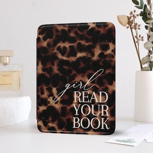 Op de afbeelding: Een rechthoekige e-reader hoes met een luipaardprint in bruin en zwart. De hoes heeft de tekst "girl READ YOUR BOOK" in witte letters. De hoes wordt getoond op een wit oppervlak, met een parfumfles en een vaas op de achtergrond.