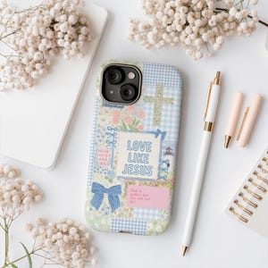 May include: A phone case with a patchwork design featuring a cross, flowers, and the text "LOVE LIKE JESUS." The case has a blue bow and the text "HOW LUCKY ARE WE." A white pen and two pink lipsticks are also visible.