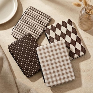 Puede incluir: Cuatro fundas rectangulares para lector electrónico con distintos patrones: una con estampado de pata de gallo marrón y blanco, una con diseño de rombos argyle, una con cuadros vichy y otra con un patrón de estrellas marrón y blanco.