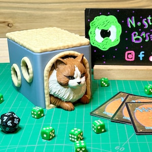 Può includere: Una casetta per gatti blu e beige con una statuetta di gatto marrone e bianco che sporge dall'apertura. La casetta ha due aperture rotonde con bordi bianchi. Dadi verdi sono sparsi su un tappetino verde davanti alla casetta. Due carte con bordi neri e testo bianco sono sul tappetino. Il testo sulle carte è "Deck" e "Magic".