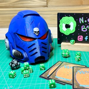 Könnte beinhalten: Ein blauer Helm mit Totenkopf-Emblem, roten Augenlinsen und einem schwarz-silbernen Design. Grüne Würfel, Spielkarten und ein Schild mit grünem Text und einem Logo sind ebenfalls vorhanden. Die Artikel befinden sich auf einer grünen Rasteroberfläche.