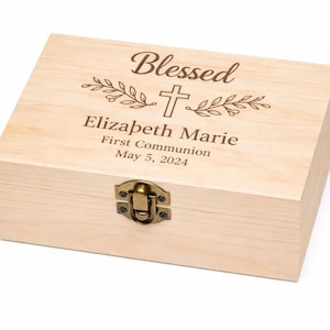 以下が含まれることがあります： 蝶番式の蓋と金属製の留め金が付いた、明るい色の木製ボックス。ボックスには「Blessed」の文字、十字架、名前「Elizabeth Marie」、そして「First Communion May 5, 2024」が刻まれています。