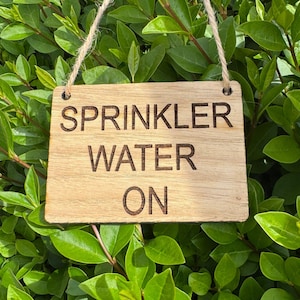 以下が含まれることがあります： 黒い文字で「SPRINKLER WATER ON」と書かれた木製の看板。看板は紐で吊り下げられており、緑の茂みの前にある。