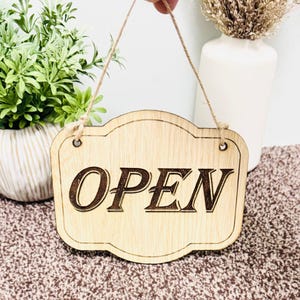 May include: A wooden sign with a rope hanger that says "OPEN" in a decorative font. The sign is light brown and has a distressed look.