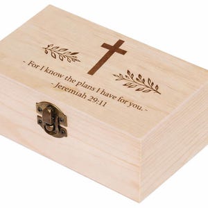 Puede incluir: Una caja de madera de color claro con tapa con bisagras y cierre metálico. La parte superior de la caja presenta una cruz marrón, diseños de hojas y el texto "For I know the plans I have for you. - Jeremiah 29:11."