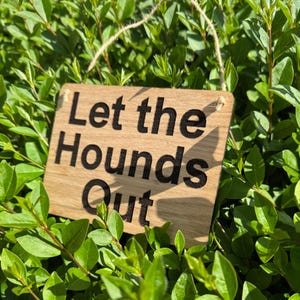 May include: A wooden sign with the text "Let the Hounds Out" in black lettering. The sign is hanging from a string and is surrounded by green foliage.