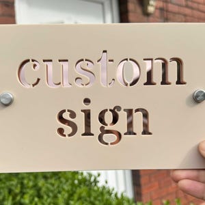 Può includere: Un cartello personalizzato beige con le parole "custom sign" tagliate in uno stile a stencil.
