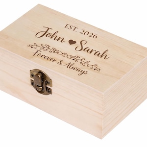 May include: A light-colored wooden box with a hinged lid and a metal clasp. The box is engraved with the names "John" and "Sarah", a heart symbol, and the words "Forever & Always" and "Est. 2026".