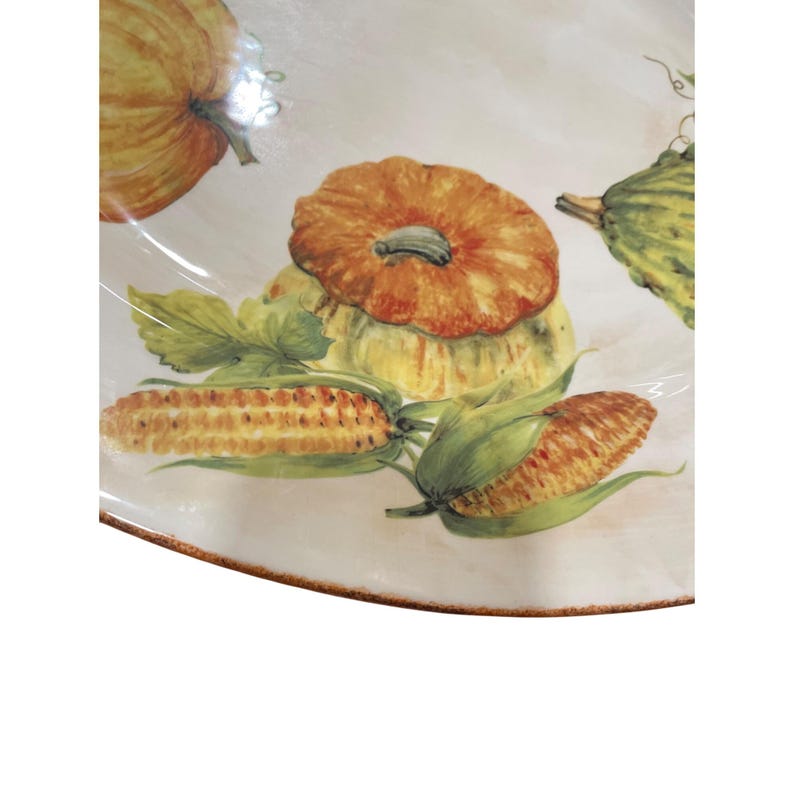 May include: A decorative plate featuring a watercolor design of pumpkins, squash, and corn on the cob. The plate has a cream-colored background with a brown rim. The pumpkins are orange, the squash is yellow and green, and the corn is yellow and brown.