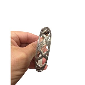 May include: Silver-toned bangle bracelet with an openwork design. The bracelet features an interwoven pattern and is embellished with small, sparkling stones. The bracelet is held in a hand against a white background.
