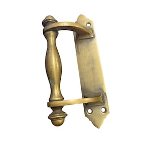 May include: Antique brass door handle with a curved handle and a rectangular backplate. The handle has a decorative, ornate design and is likely vintage. The brass has a warm, golden hue with some signs of age and wear.