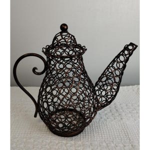May include: A decorative metal teapot-shaped object with a bronze finish. The teapot is constructed from interconnected circles, creating an openwork design. It features a curved handle, a spout, and a lid with a small knob. The teapot is designed for decorative purposes.
