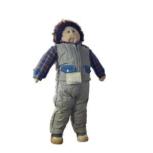 May include: A vintage doll wearing a gray quilted vest and matching pants over a plaid shirt. The doll has brown curly hair and a tag that reads "Victor Barry". The doll is wearing green and tan shoes.