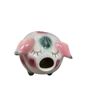 Pode incluir: Um recipiente em forma de porco de cerâmica com um design caprichoso. O porco tem orelhas cor-de-rosa, bochechas rosadas e olhos fechados com longos cílios. O recipiente é principalmente branco com detalhes em rosa, azul e vermelho escuro. O porco tem uma grande abertura.