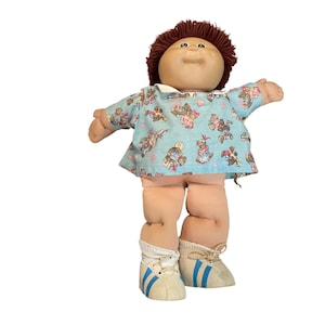 Può includere: Una bambola vintage Cabbage Patch Kid con capelli castani e carnagione chiara. La bambola indossa un top blu fantasia, pantaloncini rosa e scarpe da ginnastica bianche con strisce blu. Le braccia della bambola sono tese.