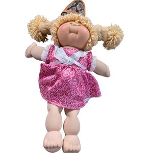 Può includere: Bambola Cabbage Patch Kid vintage con codini biondi di filo e un vestito rosa floreale. La bambola ha un viso sorridente e indossa una fascia marrone con motivo a pelle di serpente. Le braccia e le gambe della bambola sono tese.
