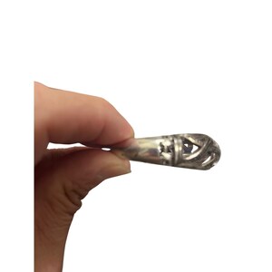 May include: A silver-colored pendant with an intricate, openwork design. The pendant has a teardrop shape with a detailed pattern of lines and cutouts. The metal has a polished finish, reflecting light. The pendant is held by a hand against a white background.