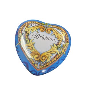 May include: A heart-shaped trinket box with a blue background and a decorative gold and white design. The box features the word "Brighton" in elegant script. The design includes floral and scroll elements.