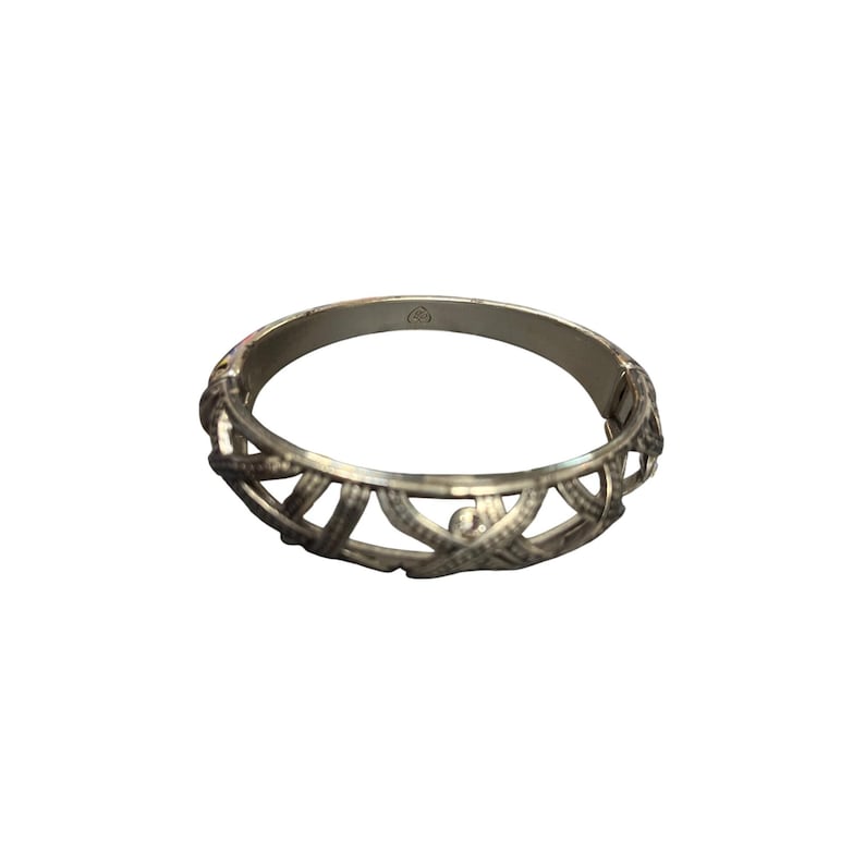 May include: Silver-toned bangle bracelet with an openwork design. The bracelet features an intricate pattern of interwoven lines, creating a textured appearance. The clasp is visible, and the bracelet is set against a white background.