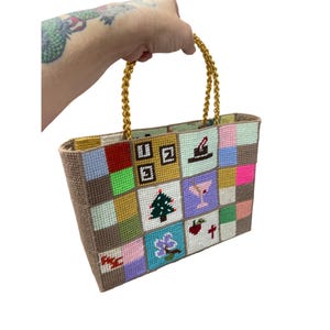 May include: A rectangular, hand-stitched tote bag with a patchwork design. The bag features a variety of colorful squares, each with a unique embroidered image. Gold chain handles complete the design. The bag is held by a person.