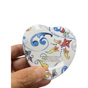 May include: Heart-shaped trinket box with a white background and a colorful floral design. The box features blue, red, yellow, and green floral patterns. Text on the box reads "Jewelry designed in California, Packaging made in China, Exclusively for BRIGHTON."