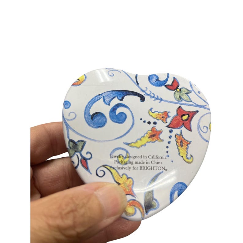 May include: Heart-shaped trinket box with a white background and a colorful floral design. The box features blue, red, yellow, and green floral patterns. Text on the box reads "Jewelry designed in California, Packaging made in China, Exclusively for BRIGHTON."