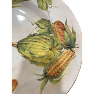 May include: A white ceramic serving dish with a hand-painted design of corn and squash. The corn is a golden yellow with green husks, and the squash is green and yellow. The rim of the dish has a distressed, aged look.