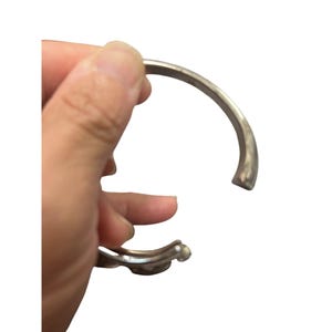 May include: Silver-toned cuff bracelet with a rounded, open design. The bracelet has a decorative element at one end, possibly a stylized floral or organic shape. The metal appears to have a slightly aged or antique finish.