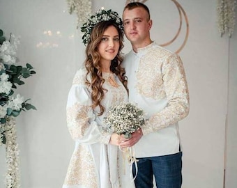 Könnte beinhalten: Ein Paar in traditioneller ukrainischer Kleidung, einem weißen bestickten Kleid und Hemd, steht zusammen in einem weißen Raum. Die Frau trägt eine Blumenkrone und hält einen Strauß weißer Blumen.
