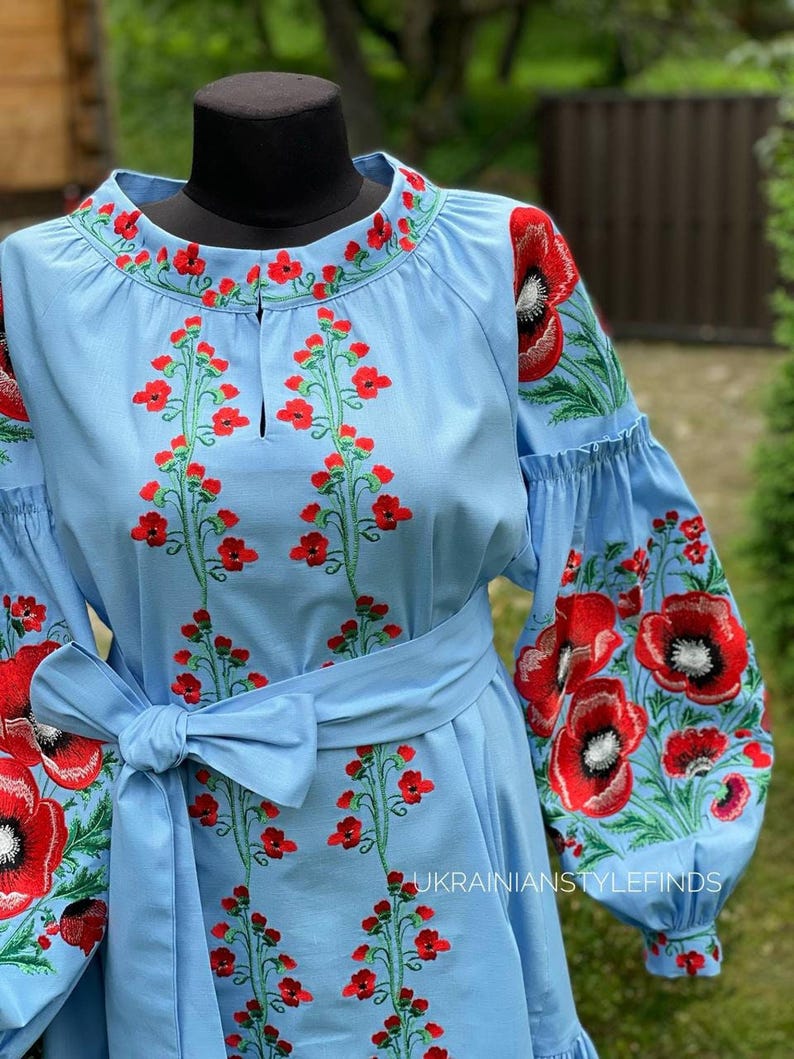 Close-up of the bodice of a light blue linen dress with red poppy embroidery on the chest and sleeves, featuring a tied belt.
