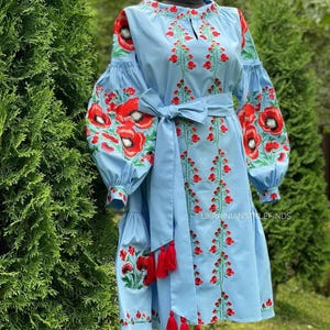 A light blue linen dress with red poppy embroidery on the sleeves and skirt, and a matching belt with red tassels, is displayed on a mannequin outdoors.