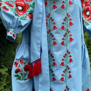 A close-up of the light blue linen dress shows the red poppy embroidery on the sleeves and skirt, with a tied belt featuring red tassels.