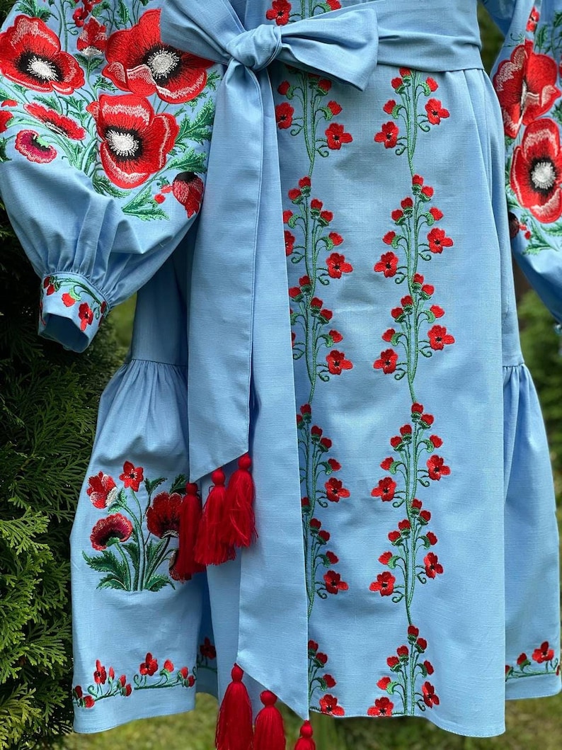 A close-up of the light blue linen dress shows the red poppy embroidery on the sleeves and skirt, with a tied belt featuring red tassels.
