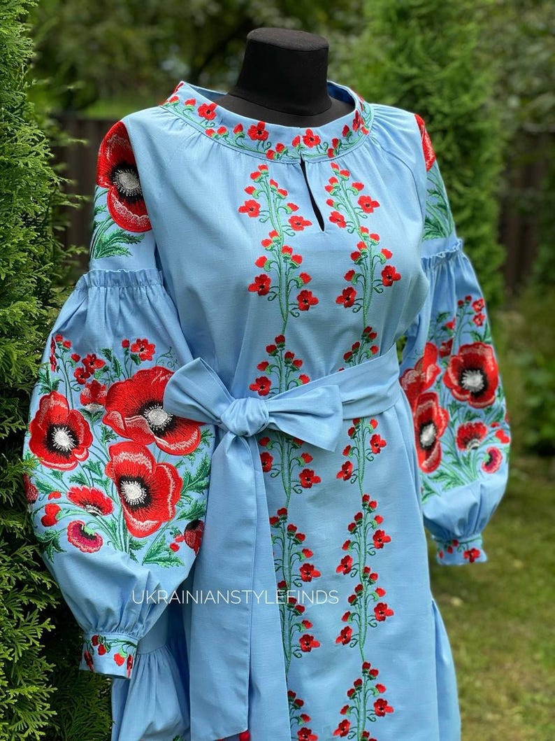 A light blue linen dress with red poppy embroidery on the bodice and sleeves, and a tied belt, is shown on a mannequin against a green hedge.