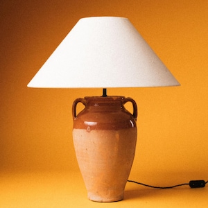 Op de afbeelding: Een keramische tafellamp met een witte lampenkap. De lampvoet is een bruin en beige vaas met twee handvatten. De lamp is aangesloten op een zwart stopcontact.