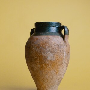 以下が含まれることがあります： 濃い緑色の縁と2つの取っ手を持つ茶色の陶器の壺。壺は素朴で古びた外観をしています。